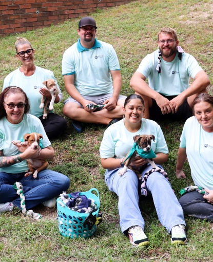 Eight people sitting on the ground, wearing teal United Way Day of Caring shirts and holding puppies.