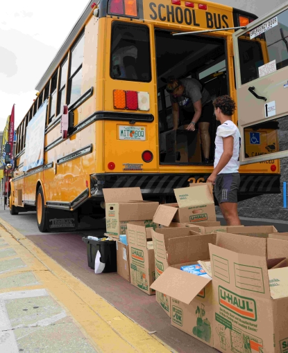 Photo of the back of a school bus opened with a teenage boy loading boxes of school supplies through the back door.