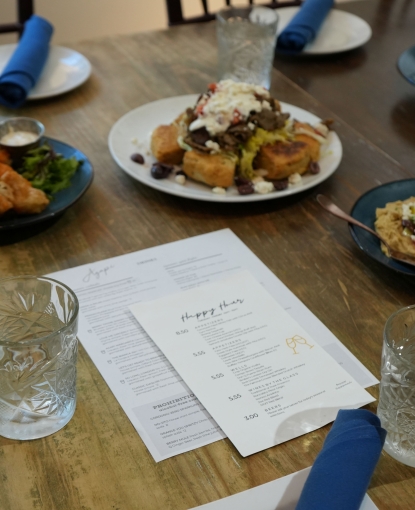 "A dinner spread at Agapi Greek cuisine, showing plated gyro appetizers, hummus dips, and menus on a wooden dining table with blue napkins and glassware.